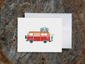 Greeting card with a red and orange camper van carrying two bikes with wood background