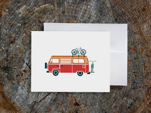 Greeting card with a red and orange camper van carrying two bikes with wood background