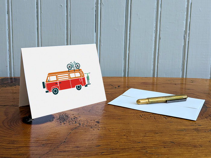 Greeting card with a red and orange camper van carrying two bikes on a table with gold pen and envelope