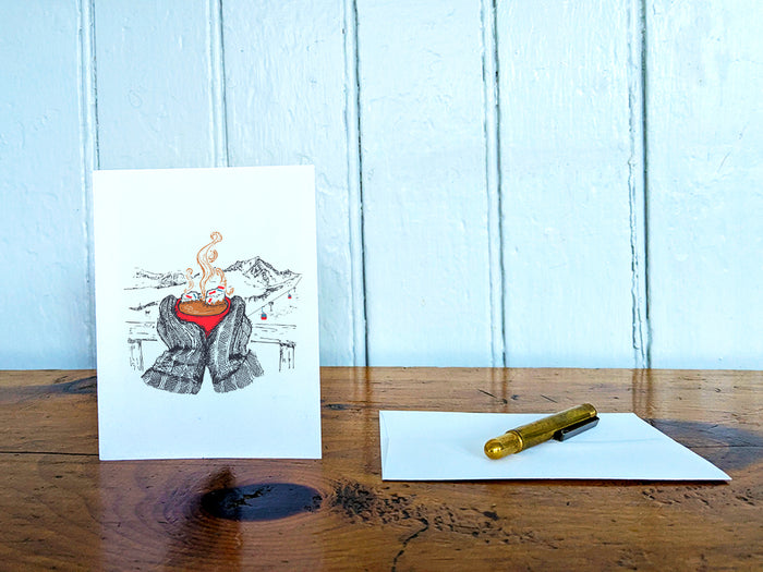 A greeting card with an illustration of a red, steaming cup of hot chocolate being held by big mittens, with mountains in the background, on a wood table next to a white envelope and gold pen.