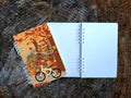 Notebook with autumn-themed cover featuring a bicycle and trees on a wooden surface