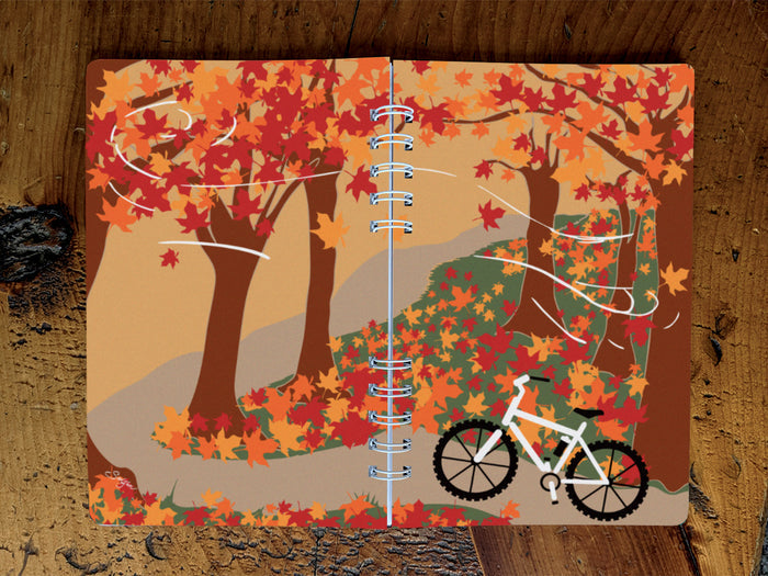 Notebook with autumn-themed cover featuring trees and a bicycle on a wooden surface