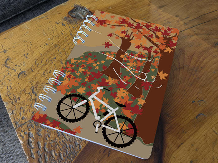 Notebook with autumn-themed cover featuring a bicycle and tree on a wooden surface