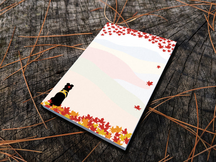 Notepad with an illustration of a black dog sitting among red, orange and yellow fall foliage with colorful rolling hills in the background, placed on a wooden surface.