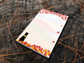 Notepad with an illustration of a black dog sitting among red, orange and yellow fall foliage with colorful rolling hills in the background, placed on a wooden surface.