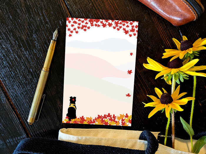 Notepad with an illustration of a black dog sitting among red, orange and yellow fall foliage with colorful rolling hills in the background, placed on a wooden surface with a gold pen, and yellow flowers.