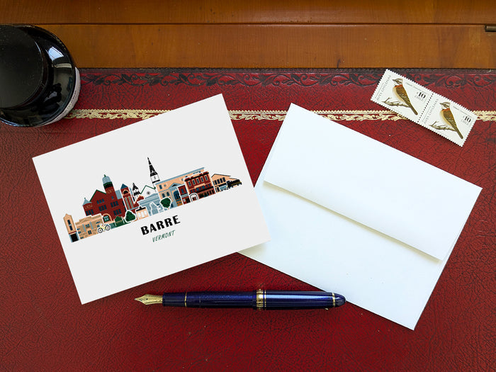 Greeting card with image of buildings arranged in a city skyline with the text Barre Vermont on a table with blue pen and envelope and stamps.