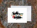 Greeting card of black dog with a red helmet sleeping with a bike and sign that reads Rest Area in the background, with a white envelope and placed on a wooden surface.