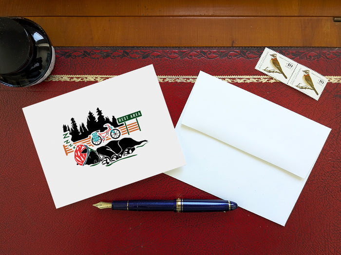 Greeting card of black dog with a red helmet sleeping with a bike and sign that reads Rest Area in the background on a table with a blue pen and envelope and stamps