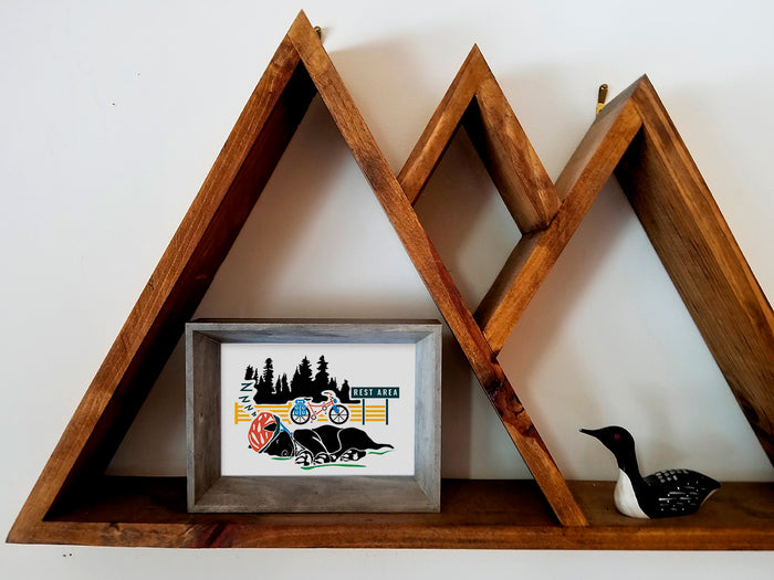 Framed art print on a wooden shelf of a black dog sleeping with a helmet on and a bike and trees in the background.
