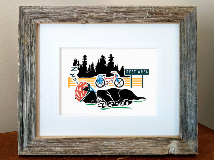 Framed art print of a black dog sleeping with a helmet on and a bike and trees in the background.