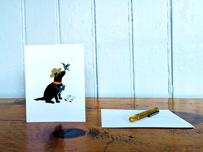 White card with drawing of a dog with binoculars and a hummingbird on a wooden surface with a pen and envelope.