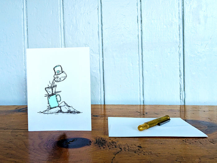 White card with a black ink drawing of pour-over coffee on a wooden surface with a pen and envelope.