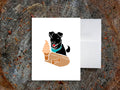 Greeting card with image of black dog looking at a soft serve ice cream on a table with a dog treat that says "creemee", with a white envelope in the background and placed on a wood surface.