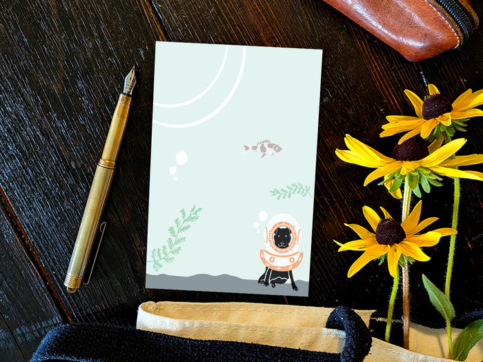 Notepad with a dog underwater with old school diving helmet and red fish, placed on a wood surface with a gold pen, and yellow flowers.