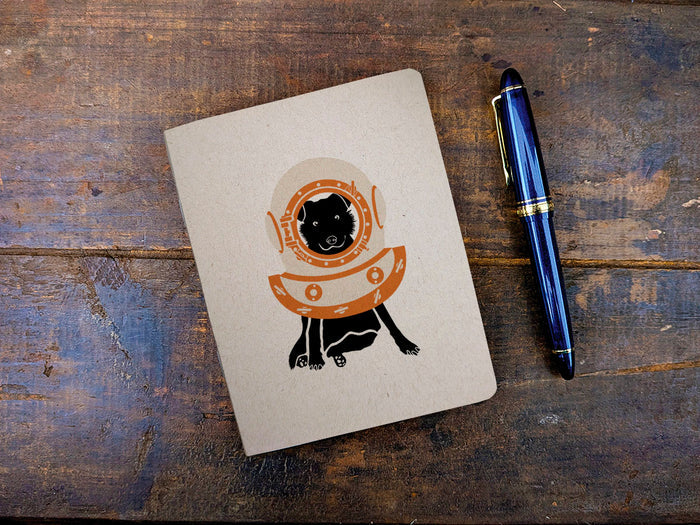 Notebook with a dog with scuba helmet illustration on a wooden surface next to a blue pen.