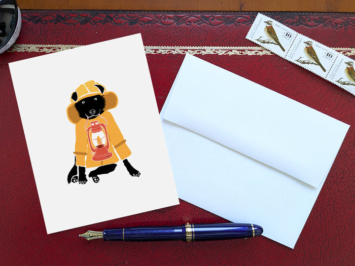 Greeting card with image of a dog with a yellow fisherman's jacket holding a lamp on a table with a blue pen and envelope and stamps.