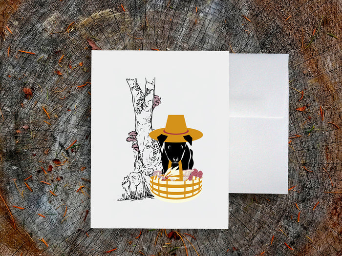 White card with drawing of dog holding basket with mushrooms on a wooden surface.