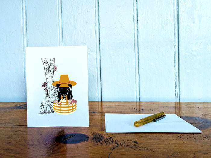 White card with drawing of dog holding basket with mushrooms on a wooden surface with a pen and envelope.