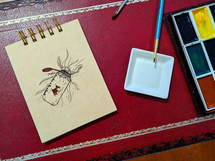 Notebook with a drawing a water bottle with a gold fish, next to a paint palette and brush on a red surface.