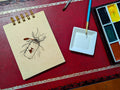 Notebook with a drawing a water bottle with a gold fish, next to a paint palette and brush on a red surface.