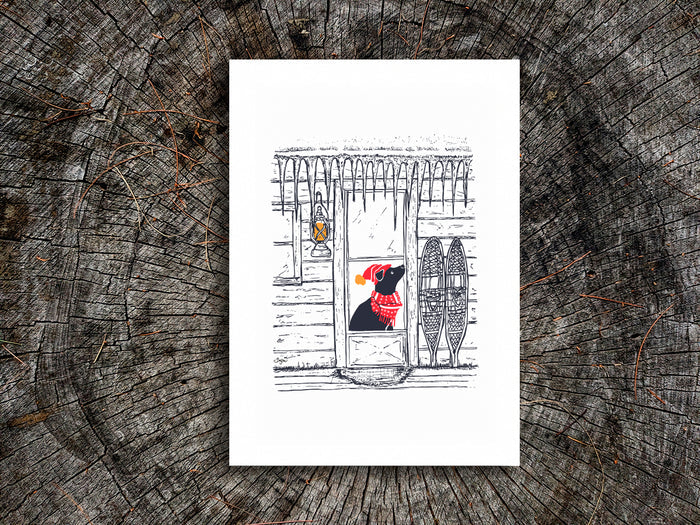 Art print featuring an illustration of a dog with red scarf and hat in a cabin covered in icicles, displayed on a wooden surface.