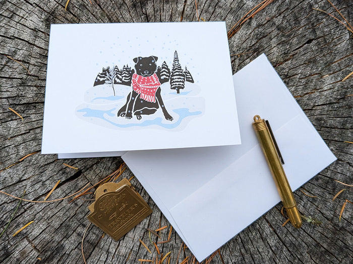 Greeting card with image of black dog with red scarf among a snowy winter landscape.