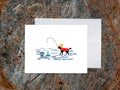 Greeting card with image of a dog with a winter hat and sweater ice fishing with a white envelope in the background and placed on a wooden surface.