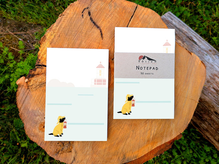Two notepads with an illustration of a dog in yellow hat and jacket holding a red lantern with a lighthouse in the background, placed on a wood surface.