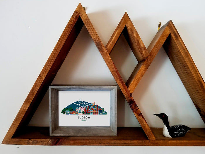 Framed art print on a wooden shelf of buildings arranged in a skyline with the text Ludlow Vermont