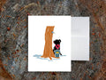 Greeting card with image of dog next to a maple syrup tap on a tree and bucket of maple syrup, with a white envelope in the background and placed on a wooden surface.