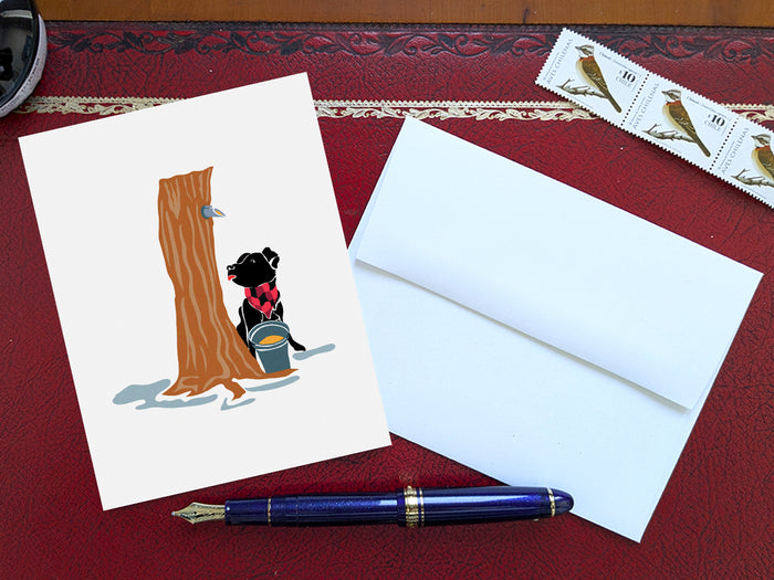 Greeting card with image of dog next to a maple syrup tap on a tree and bucket of maple syrup on a table with blue pen and envelope and stamps.