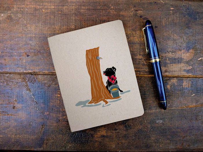 Notebook with an illustration of dog with maple syrup tap next to a blue pen on a wooden surface