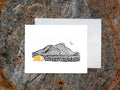Greeting card with an image of an ink illustrated mountain and bright yellow tent under a starry sky, with a white envelope in the background and placed on a wooden surface.