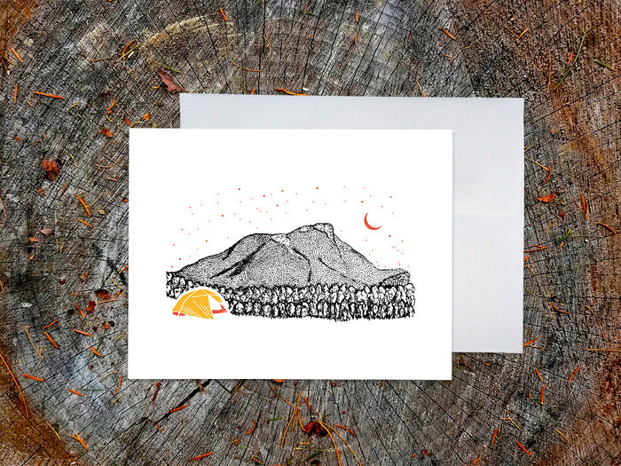 Greeting card with an image of an ink illustrated mountain and bright yellow tent under a starry sky, with a white envelope in the background and placed on a wooden surface.