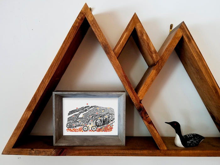 Framed art print with bicycle, leaves, and mountain illustration displayed on shelf