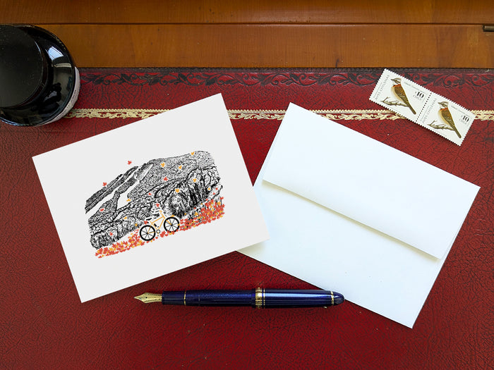 Greeting card with image of a mountain bike surrounded by brightly colored leaves with a  mountain in the background on a table with blue pen and envelope and stamps.