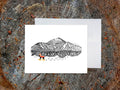 Greeting card with image of an ink illustrated mountain with ski trails and dog wearing a winter hat, vest and snowshoes, with a white envelope in the background and placed on a wooden surface.