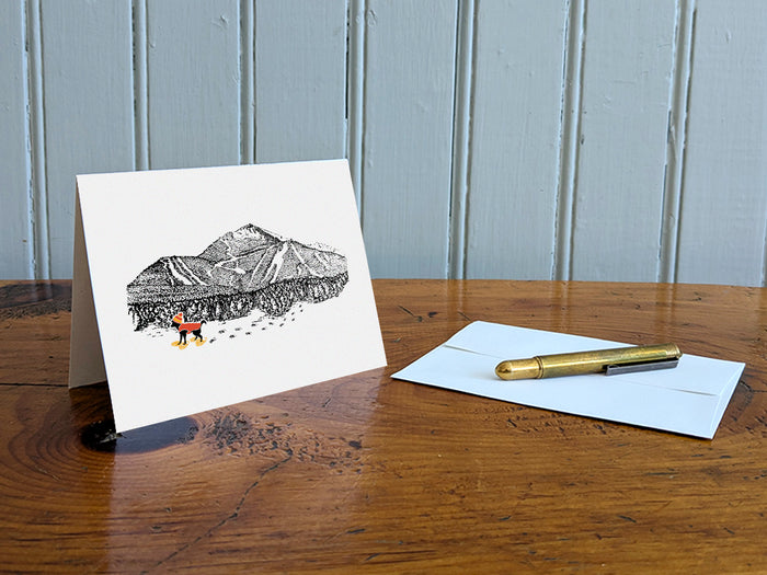 Greeting card with image of a mountain with ski trails and dog snowshoeing on a table with gold pen and envelope.