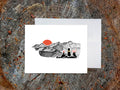 Greeting card with image of black dog looking at a red sunset behind ink illustrated mountains with envelope behind placed on a wood surface.