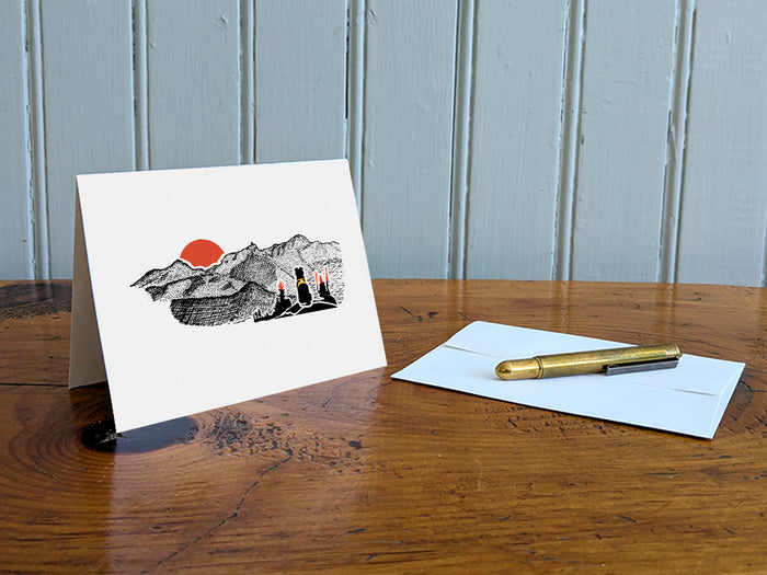 Greeting card with image of black dog looking at a red sunset behind mountains on a table with gold pen and envelope.