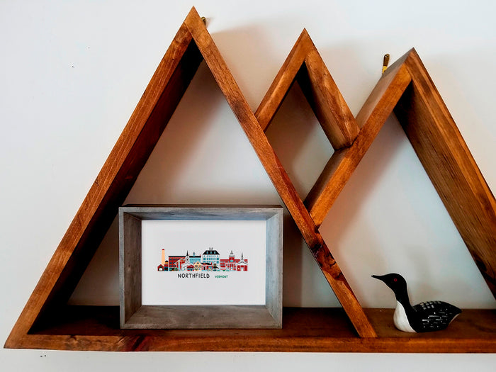 Framed art print on a wooden shelf of buildings arranged in a skyline with the text Northfield Vermont