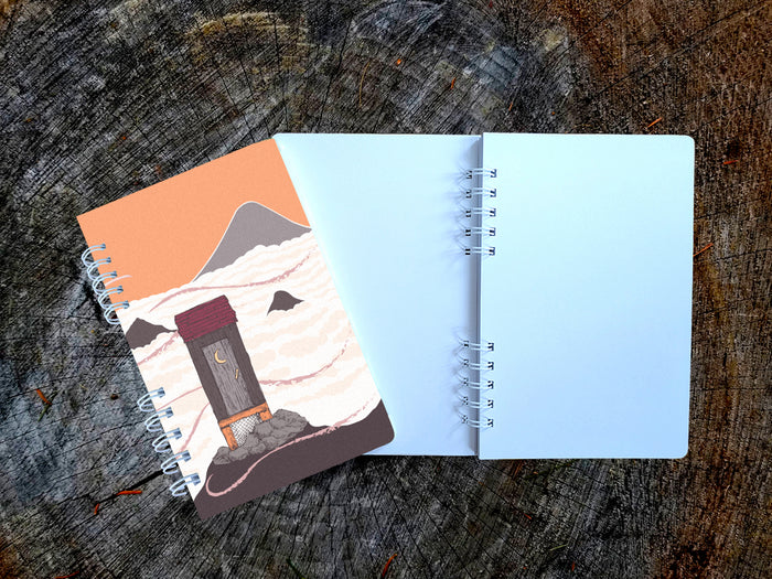 Notebook with cover featuring a mountain-top outhouse illustration on a wooden surface.