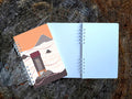 Notebook with cover featuring a mountain-top outhouse illustration on a wooden surface.