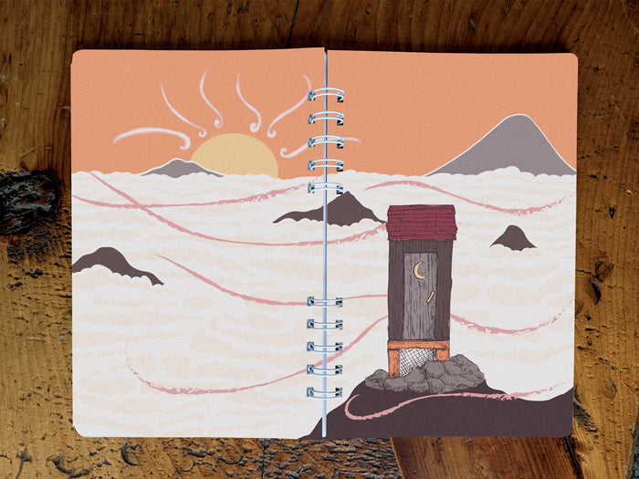 Notebook with cover featuring a mountain-top outhouse illustration on a wooden surface.