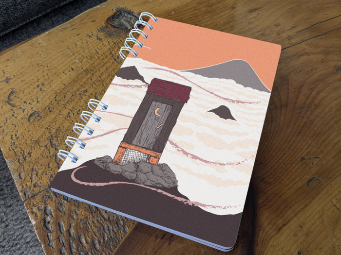 Notebook with cover featuring a mountain-top outhouse illustration on a wooden surface.