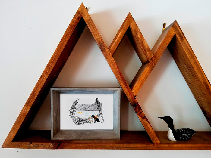 Framed illustration of a dog painting in front of a lake on a wood shelf.