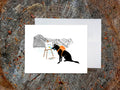 Greeting card with illustration of black dog painting in the mountains, with a white envelope placed on a wooden surface.