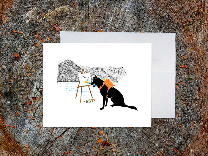 Greeting card with illustration of black dog painting in the mountains, with a white envelope placed on a wooden surface.