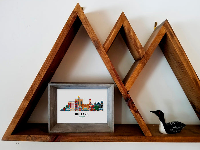 Framed art print on a wooden shelf of buildings arranged in a skyline with the text Rutland Vermont